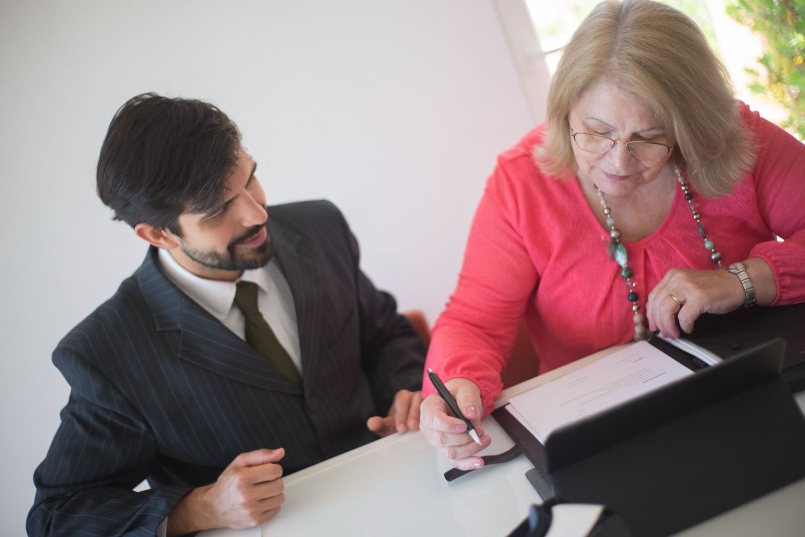 At a table, a woman in a pink blouse and a man in a suit look over a document together.