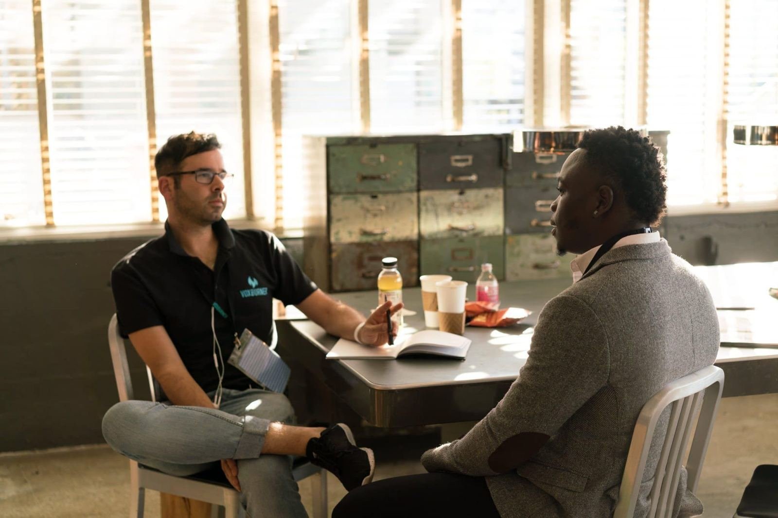 Two men sit at a table in a bright room, one gesturing while speaking to the other