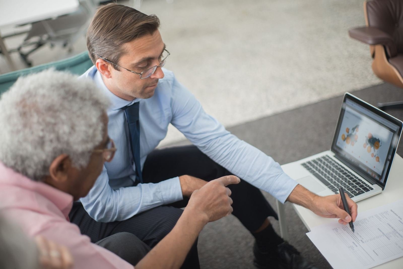 A man wearing a blue shirt and tie points at a paper while speaking with an older gentleman