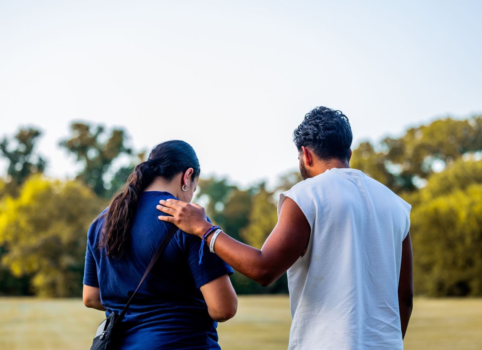 One person puts a hand on the other's shoulder as they stroll through a park.