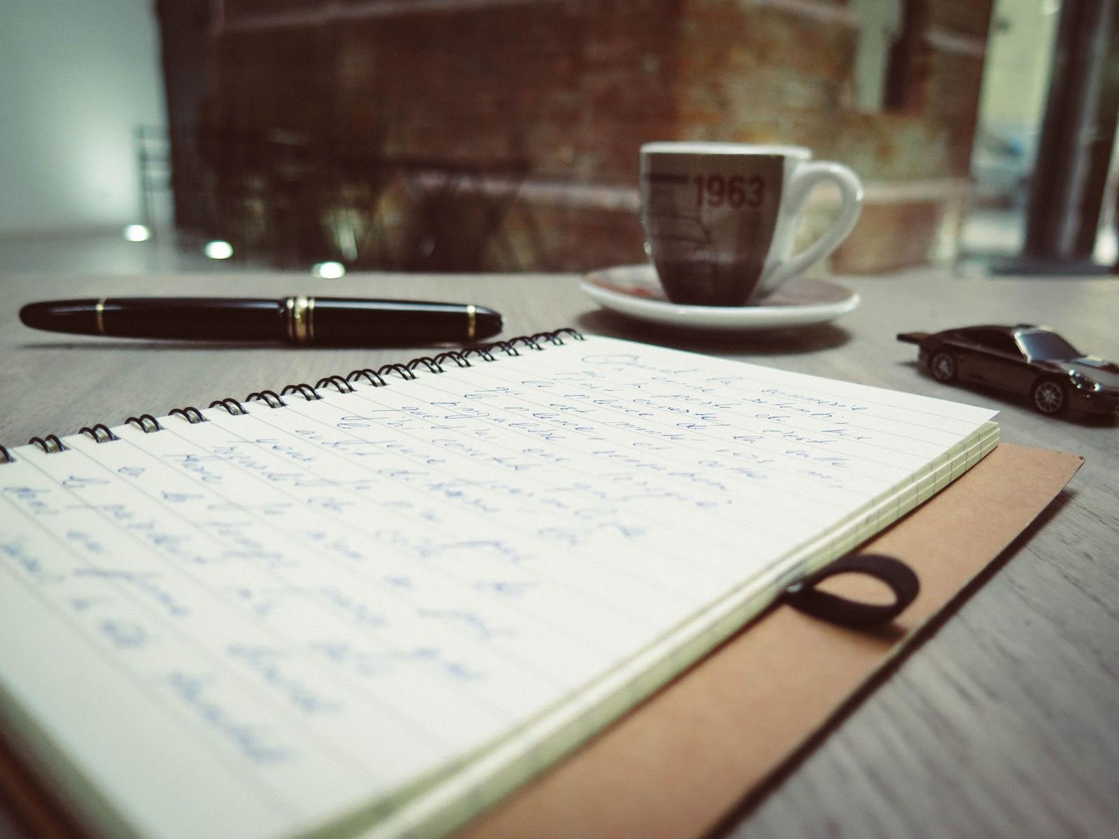 An open spiral notebook with handwritten notes, a pen, and a coffee cup on a table