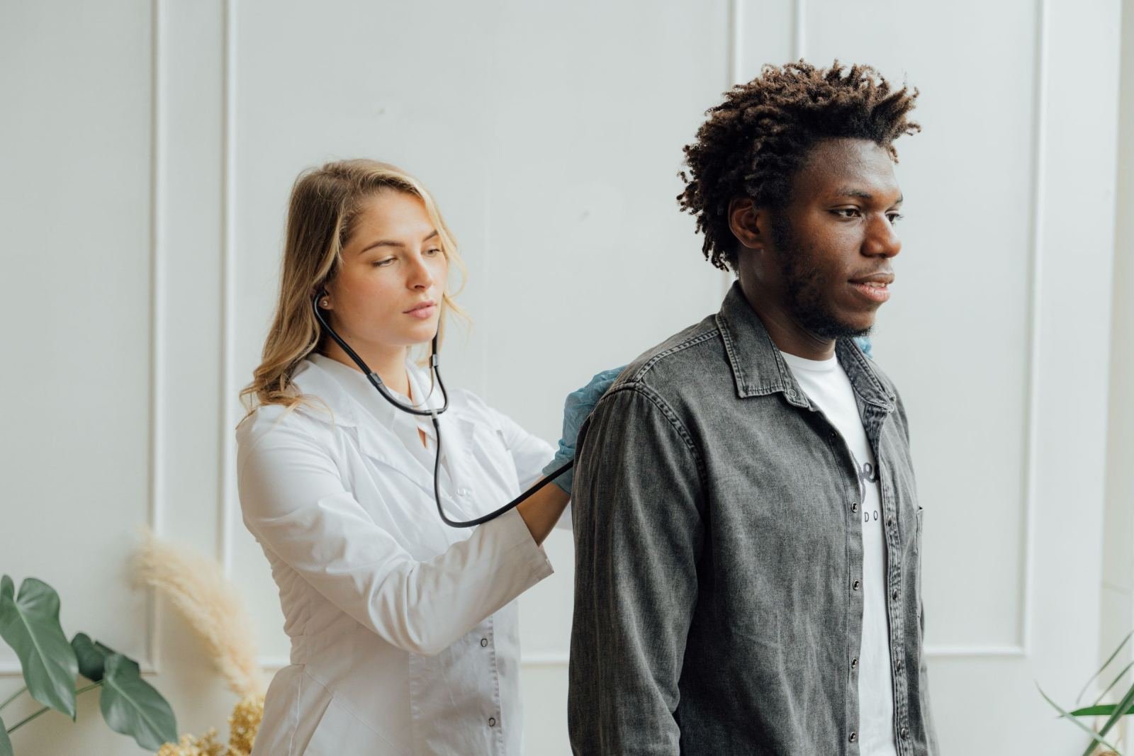 A clinician in a white coat uses a stethoscope to examine a man's back