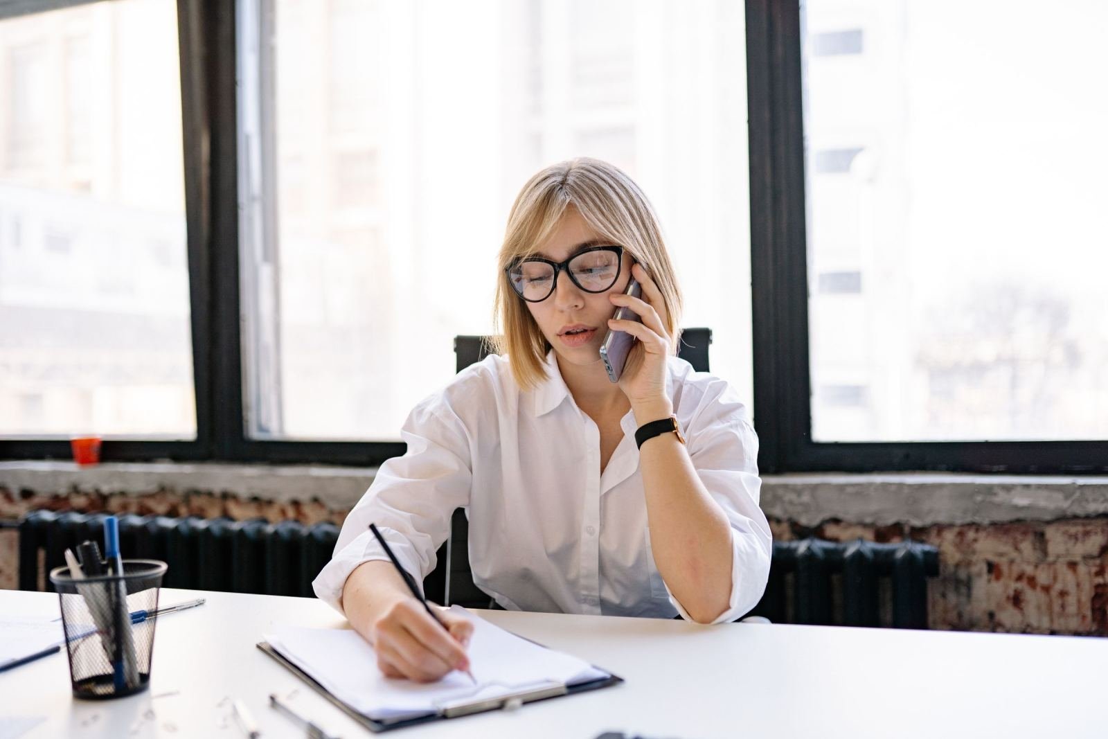 A woman with glasses talks on a mobile phone while writing in a notebook at a desk