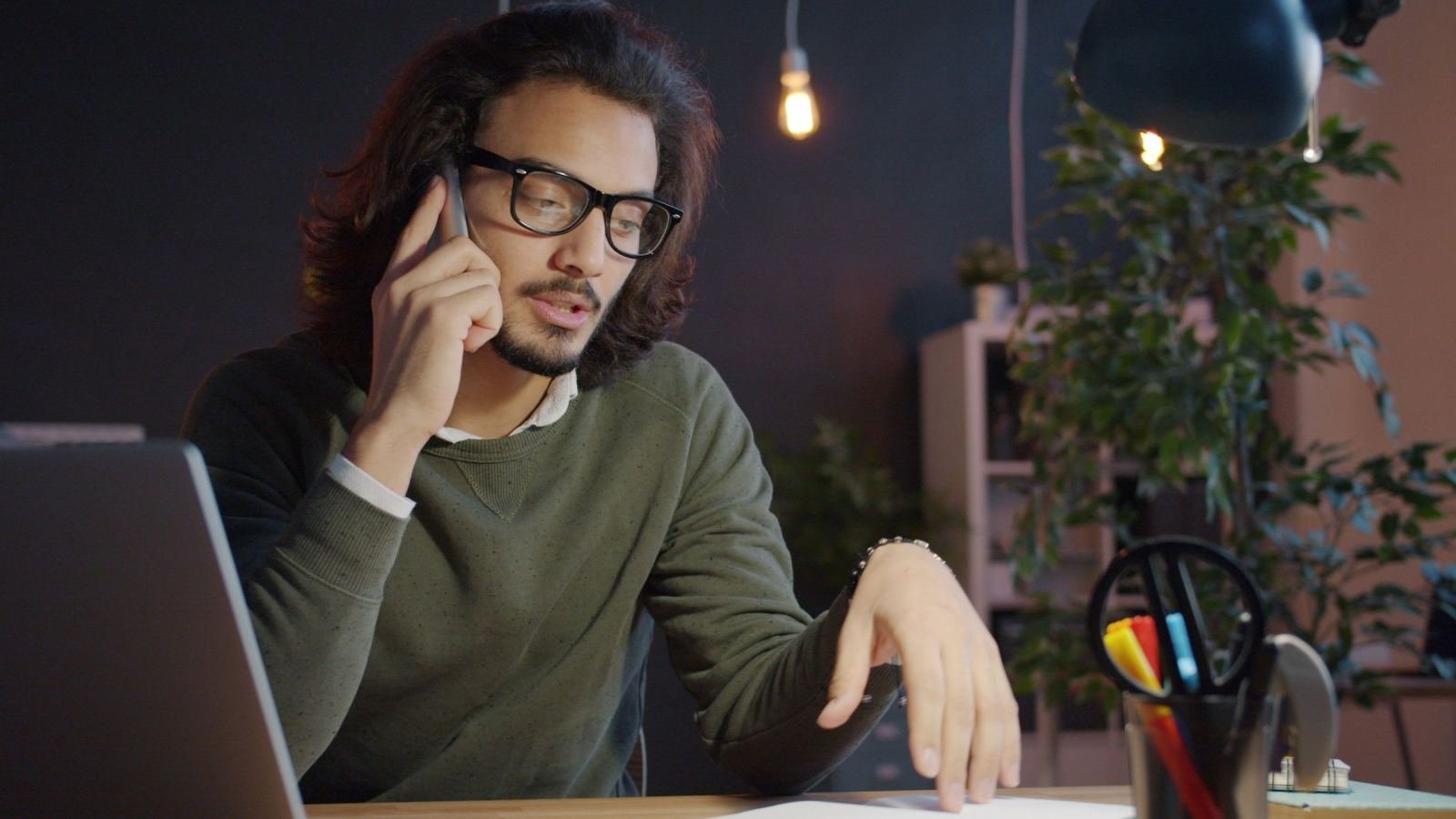 A man with glasses talks on the phone while looking at a notepad at his desk