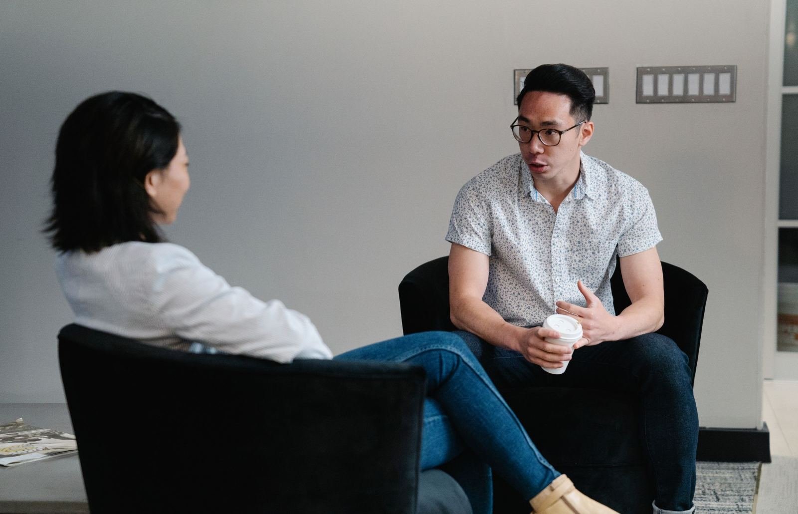 A man and woman sit in black armchairs having a conversation in a bright office