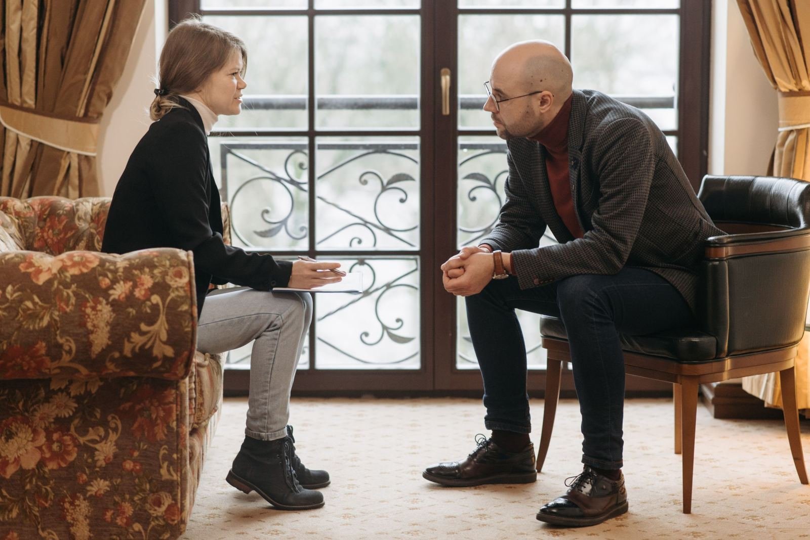 A woman and a man in glasses sit in armchairs having a conversation in a bright room
