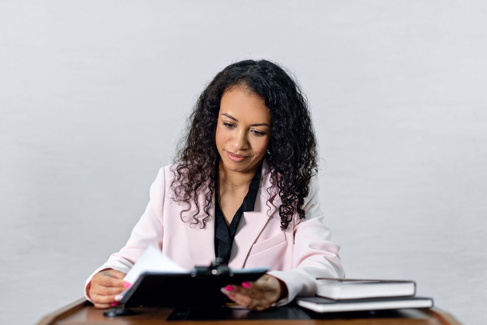 A woman in a pink blazer reads a black binder at a wooden desk