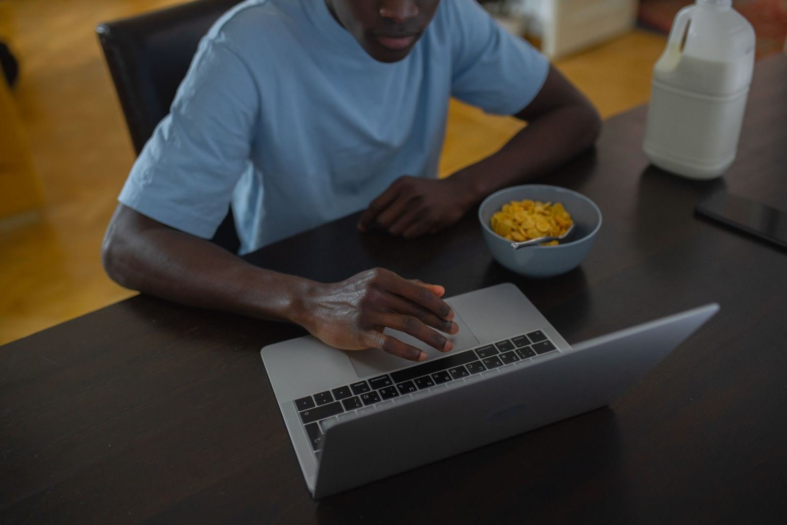 A man uses a laptop at a kitchen table with a bowl of cereal and milk