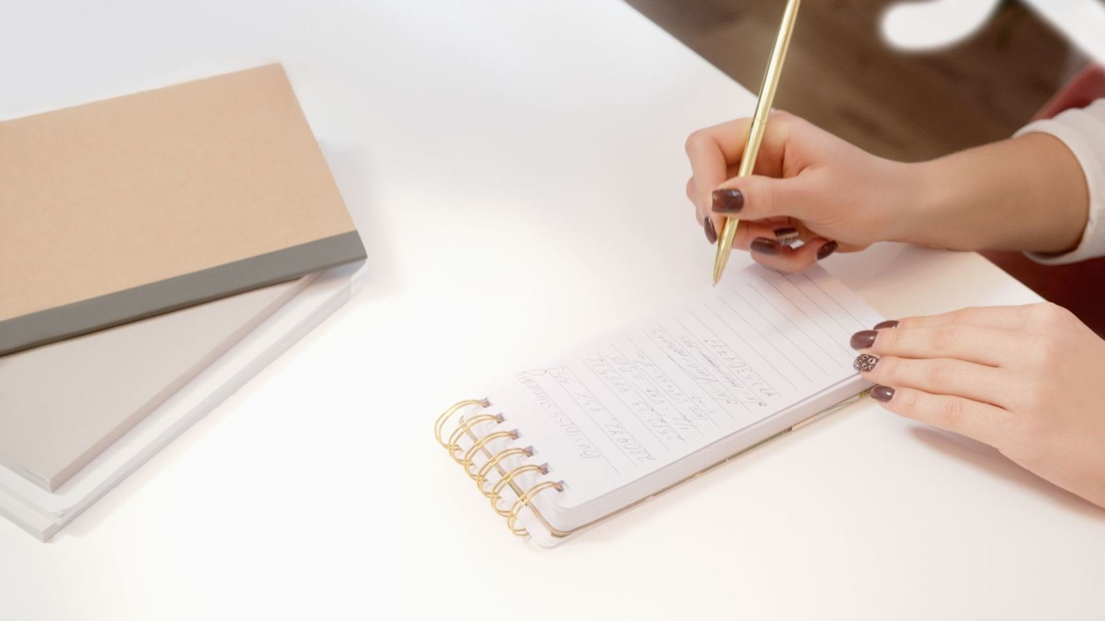 A person writes notes in a spiral notebook with a gold pen on a white desk