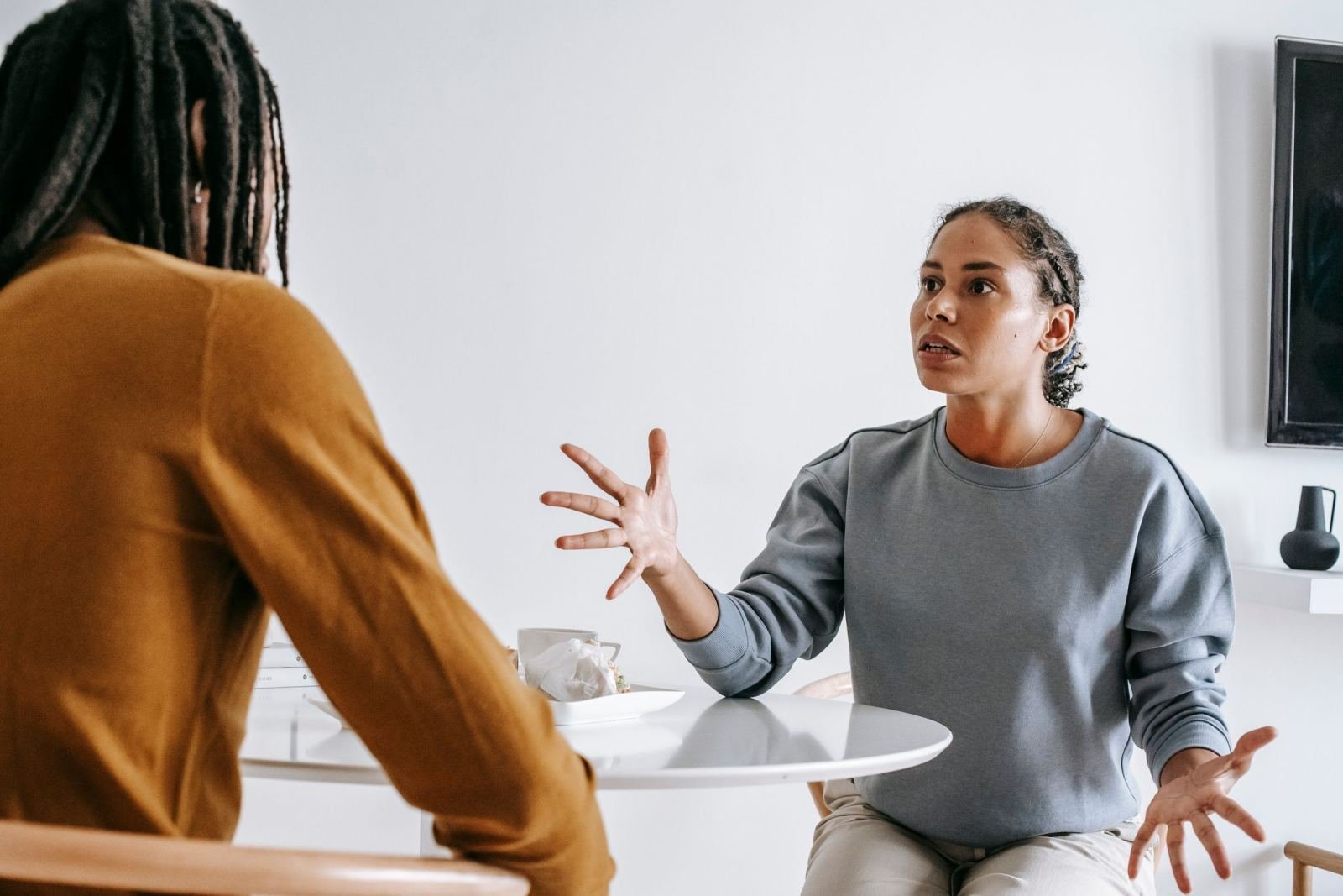 A woman in a grey sweatshirt gestures while talking to another person at a white table