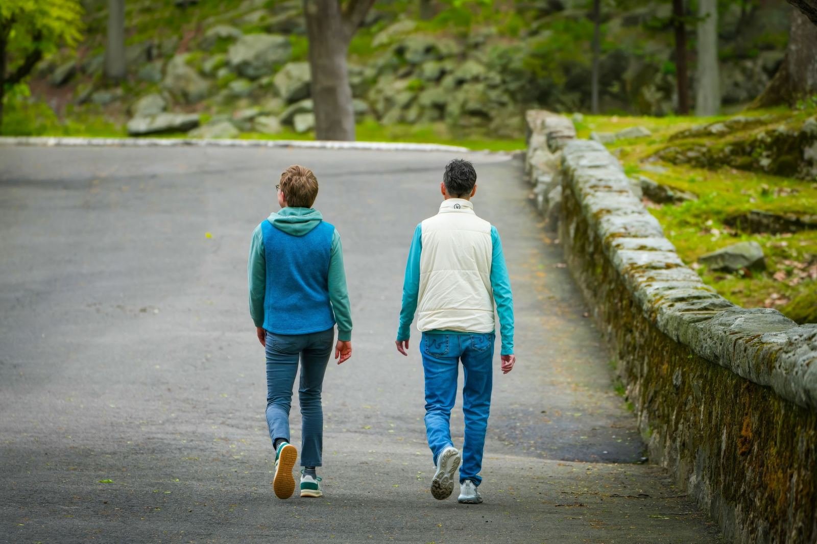 A stone wall lines a paved path where two people walk away from the camera