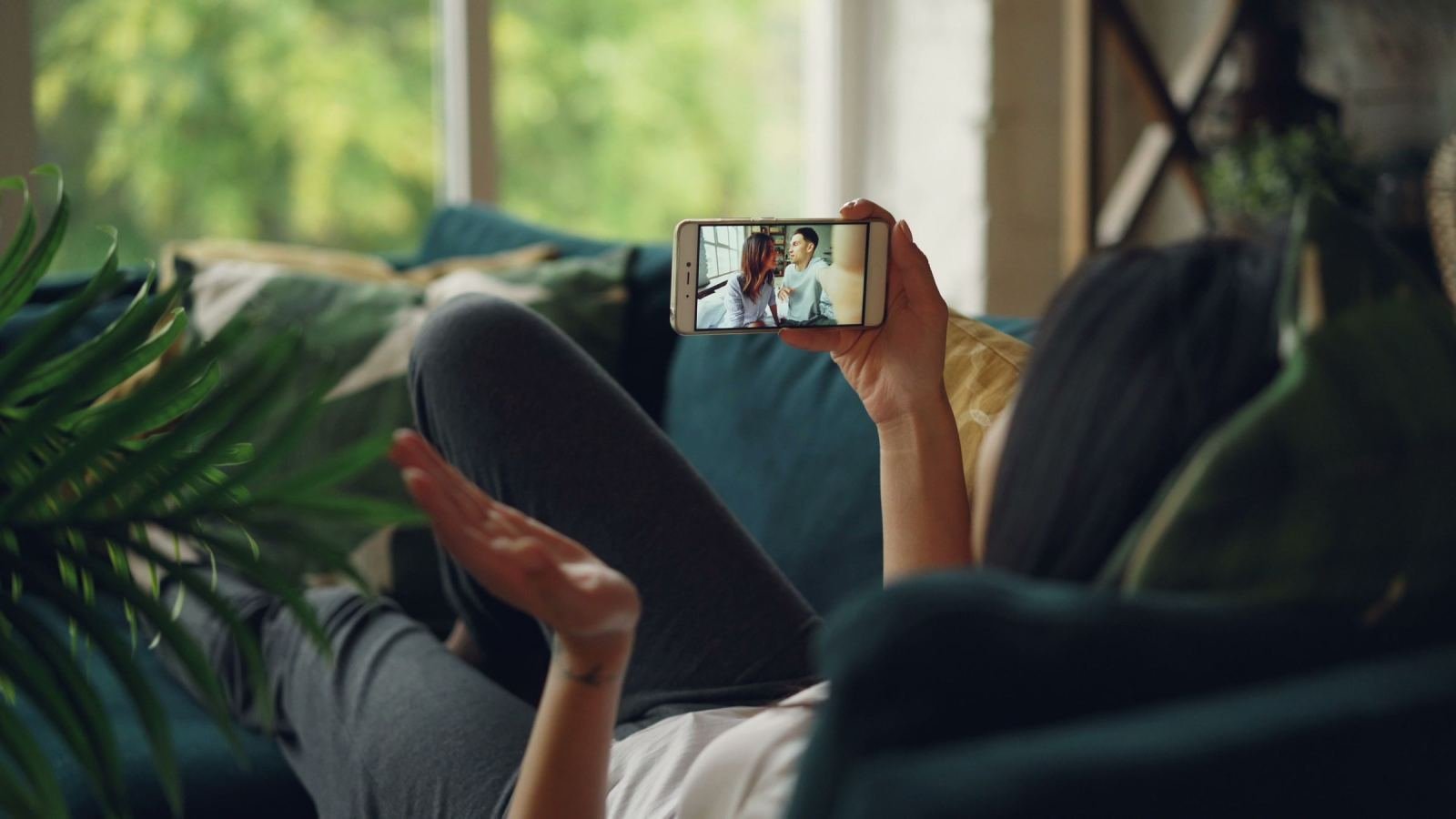 A person lying on a blue sofa holding a smartphone showing a video of two people talking
