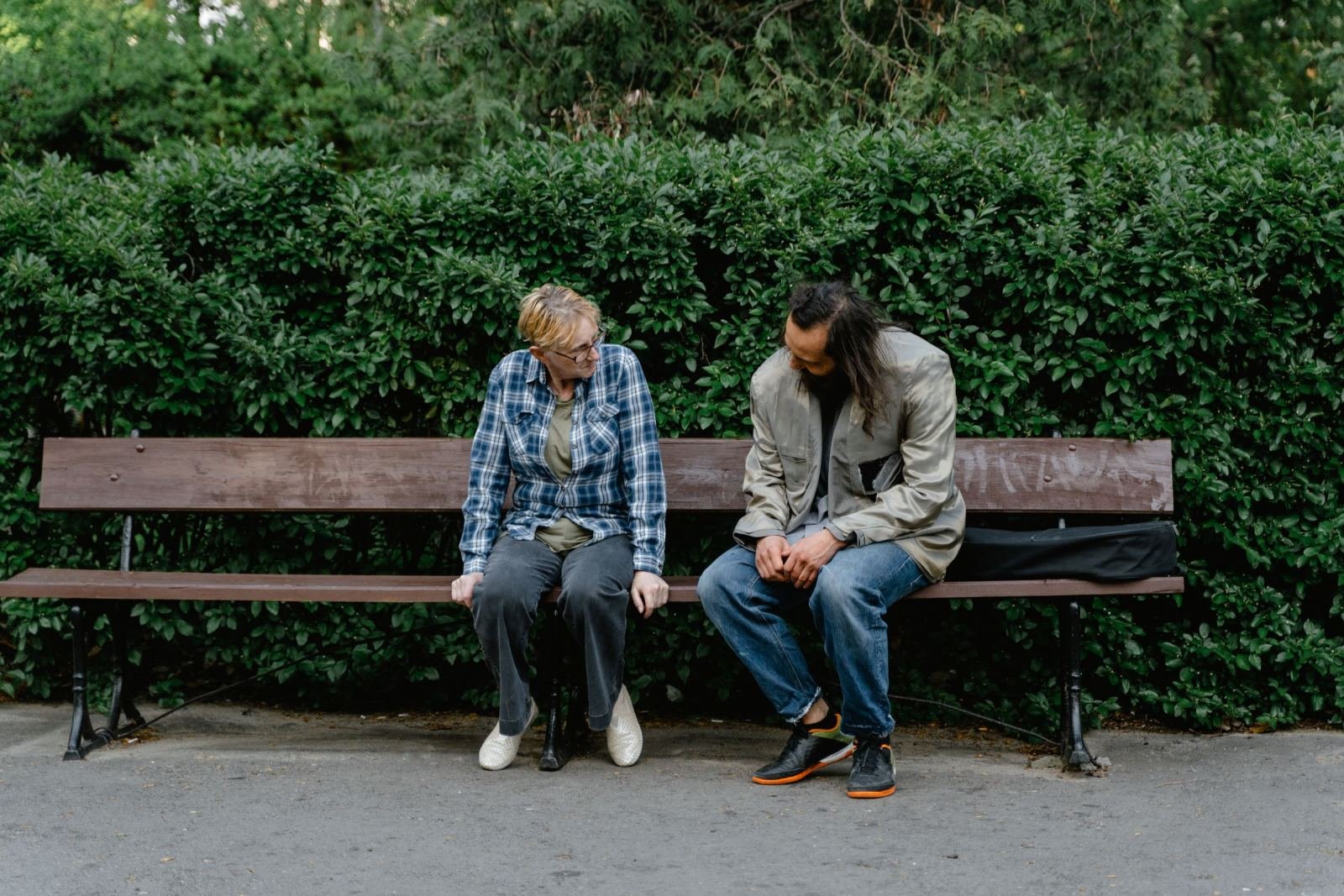 A man and woman talk on a wooden bench near a green hedge.