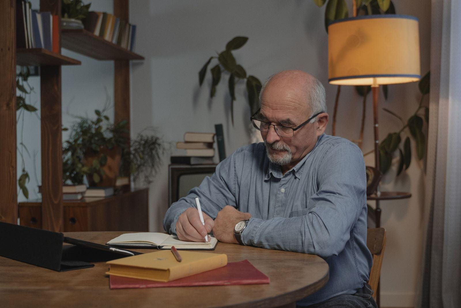 A man with glasses sits at a wooden table, writing in a notebook.