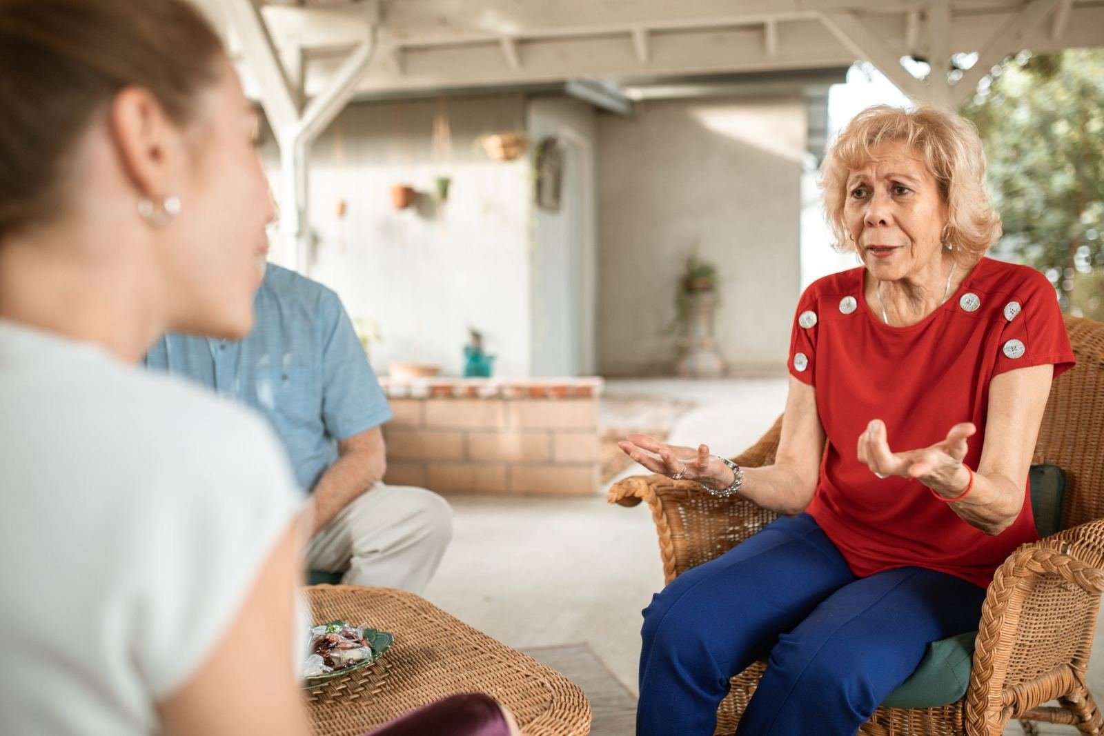 An older woman in a red shirt talks and gestures while sitting in a wicker chair on a porch