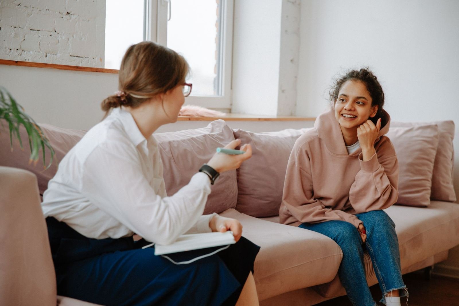 A woman in a white shirt with a notebook talks to a smiling woman in a pink hoodie