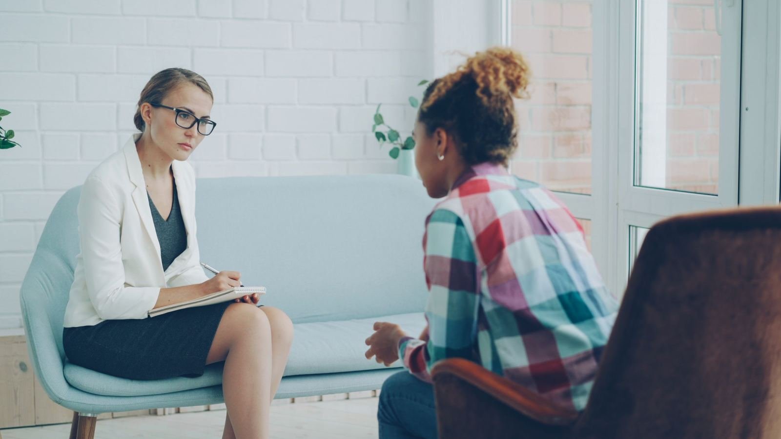 A client in a plaid shirt sits with a counselor in a white blazer. The counselor takes notes while they talk.