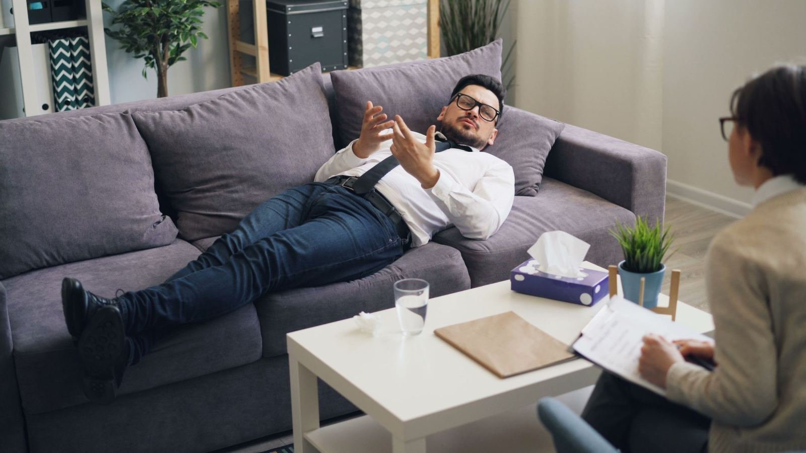 A counselor with a clipboard listens as a man rests on a couch.