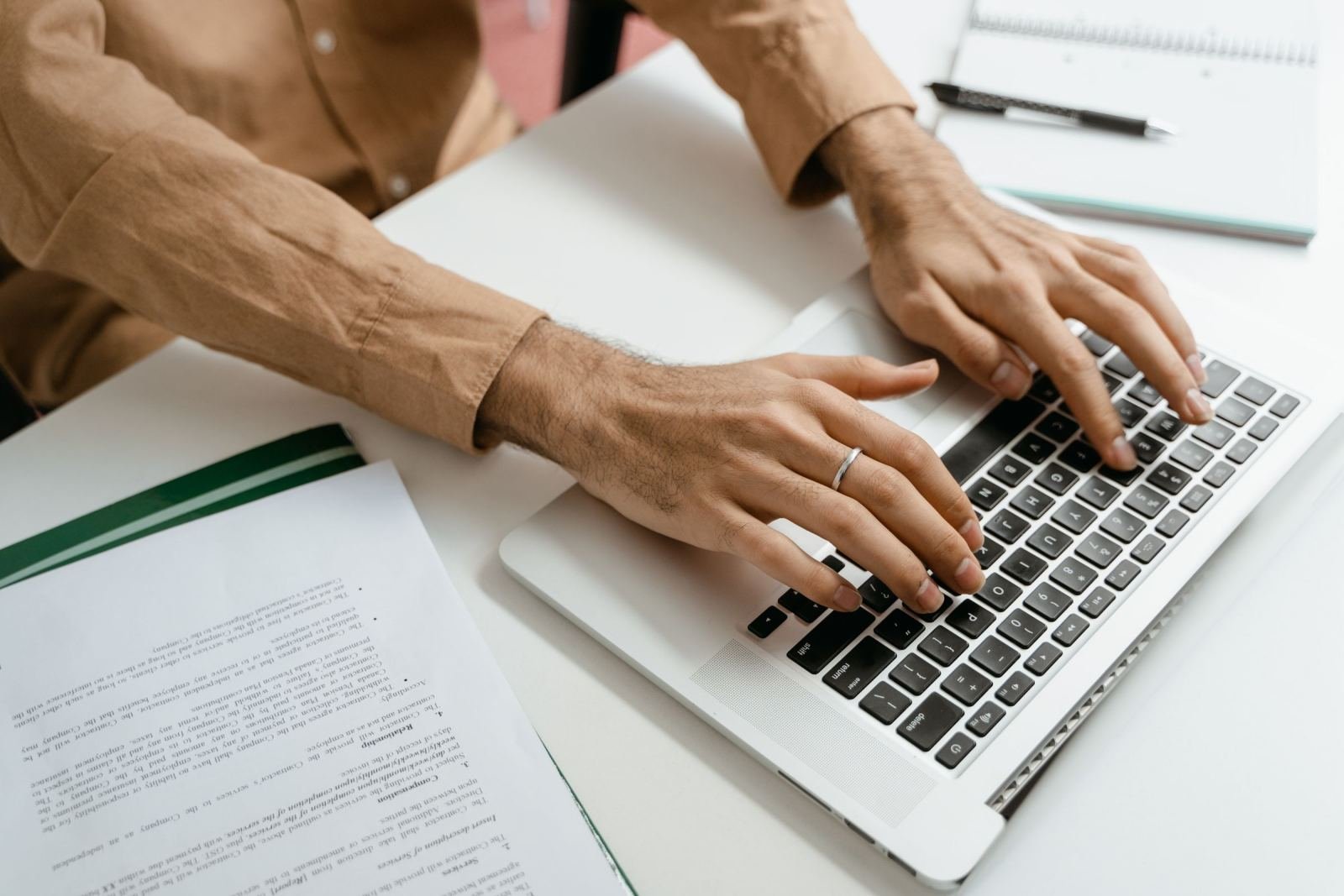 A person using a laptop at a white desk with a printed document and notebook