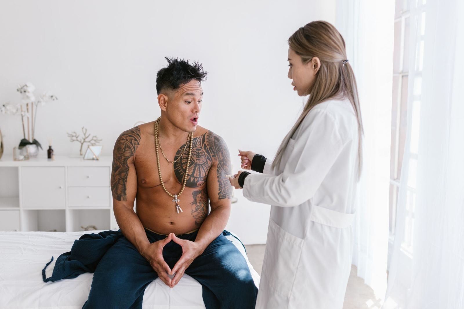 A man with tattoos sits on an exam table while a clinician in a white coat speaks with him