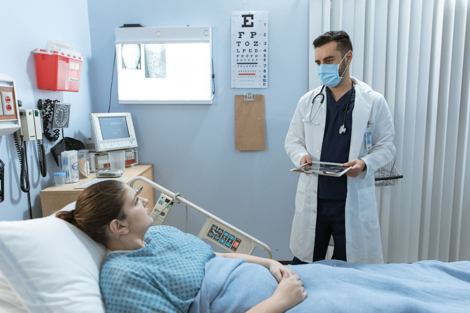 A doctor in a mask and white coat holds a tablet while speaking to a patient in a hospital bed
