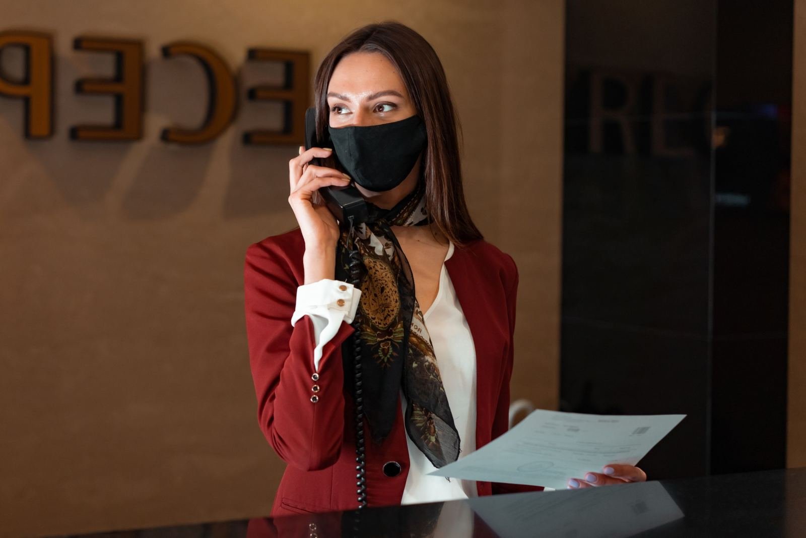 A woman in a red blazer and face mask talks on a phone at a reception desk