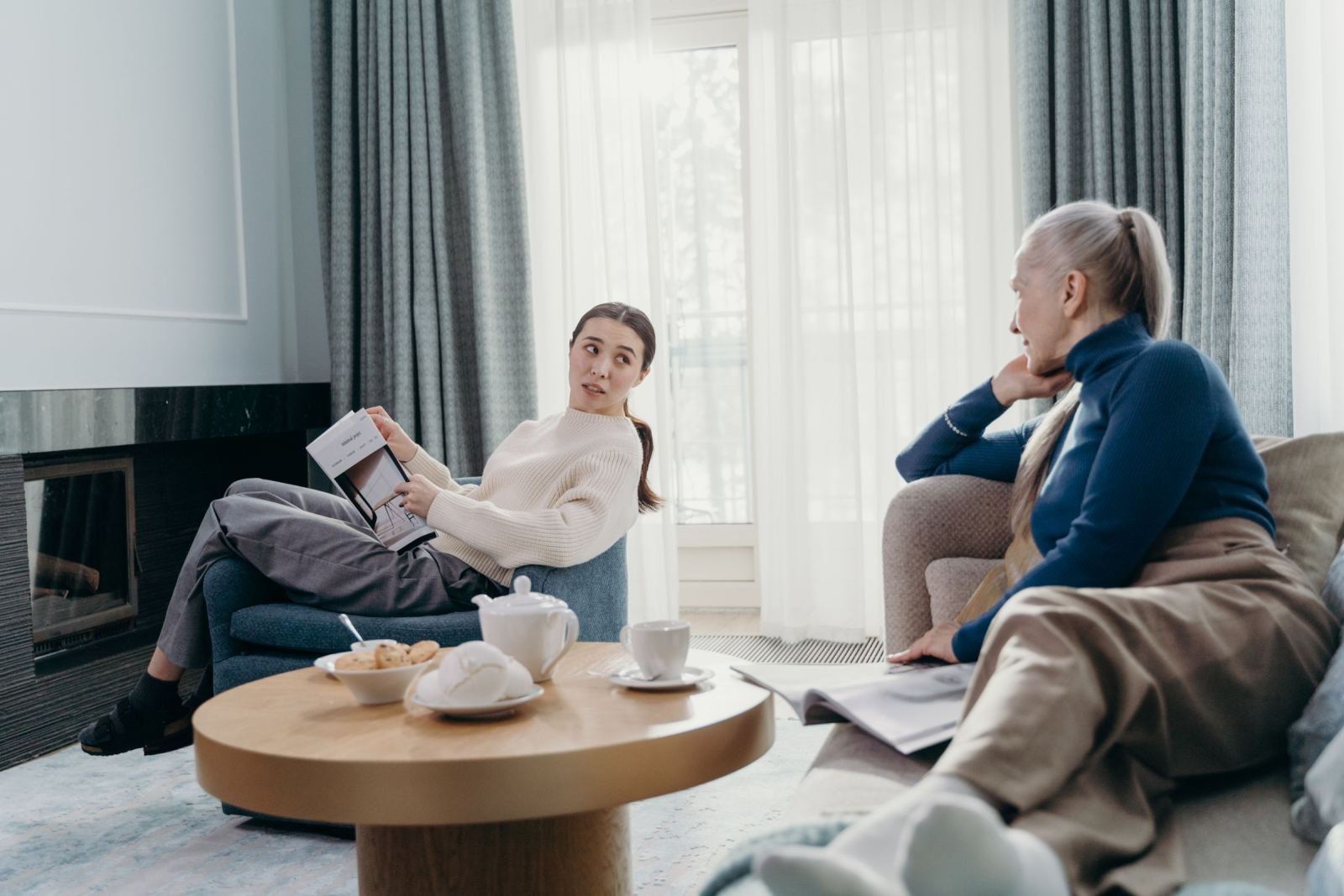 An older woman sits on a couch talking with a younger woman. A coffee table is between them in the living room.
