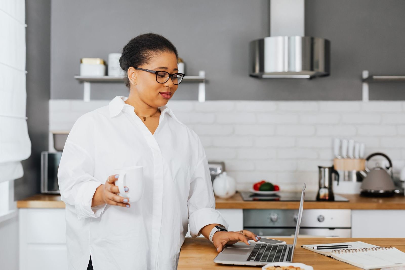 A woman in a white shirt sits in her kitchen. She holds a mug while using a laptop.
