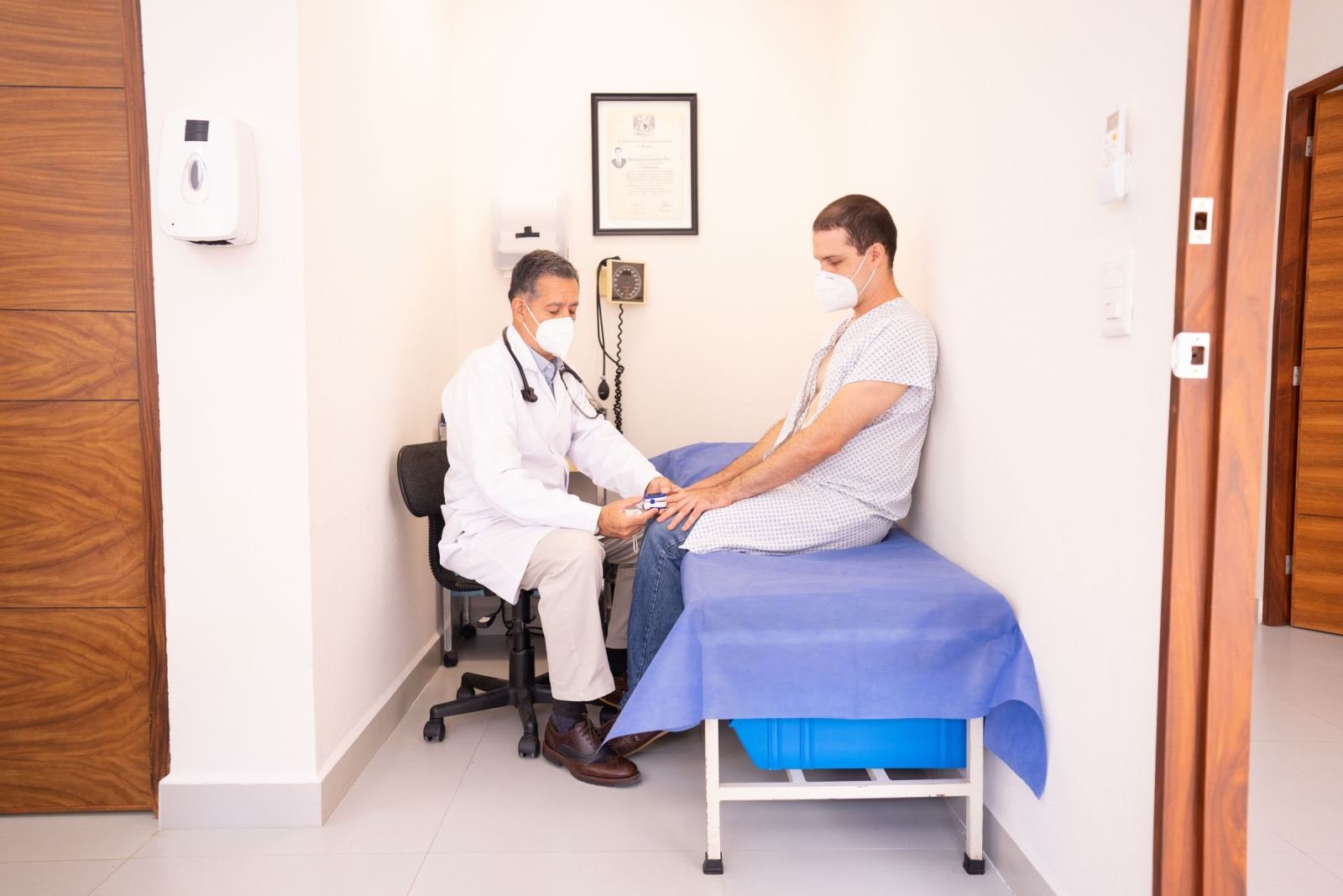 A clinician in a white coat and mask talks with a patient on an examination table