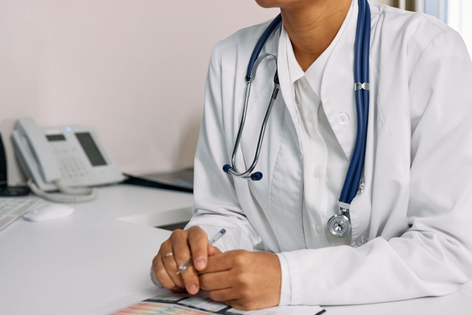 A doctor in a white coat and stethoscope sits at a desk with a pen