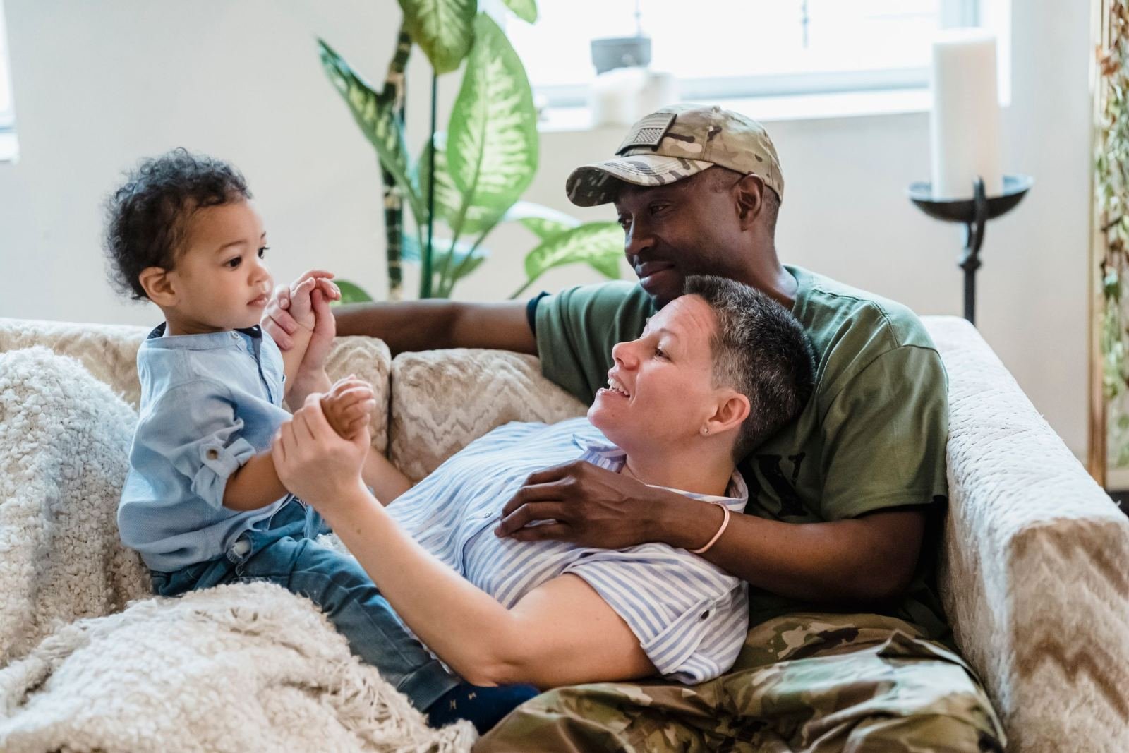 A man in a camouflage cap and a woman smile while playing with a small child on a couch