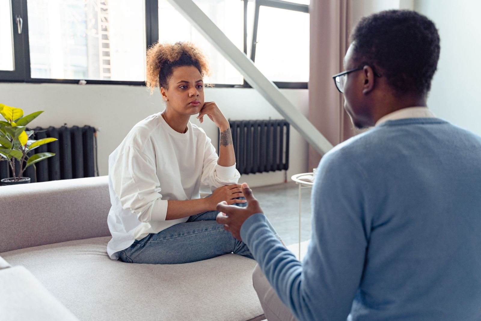 A woman sits on a sofa listening to a man who is gesturing as he speaks