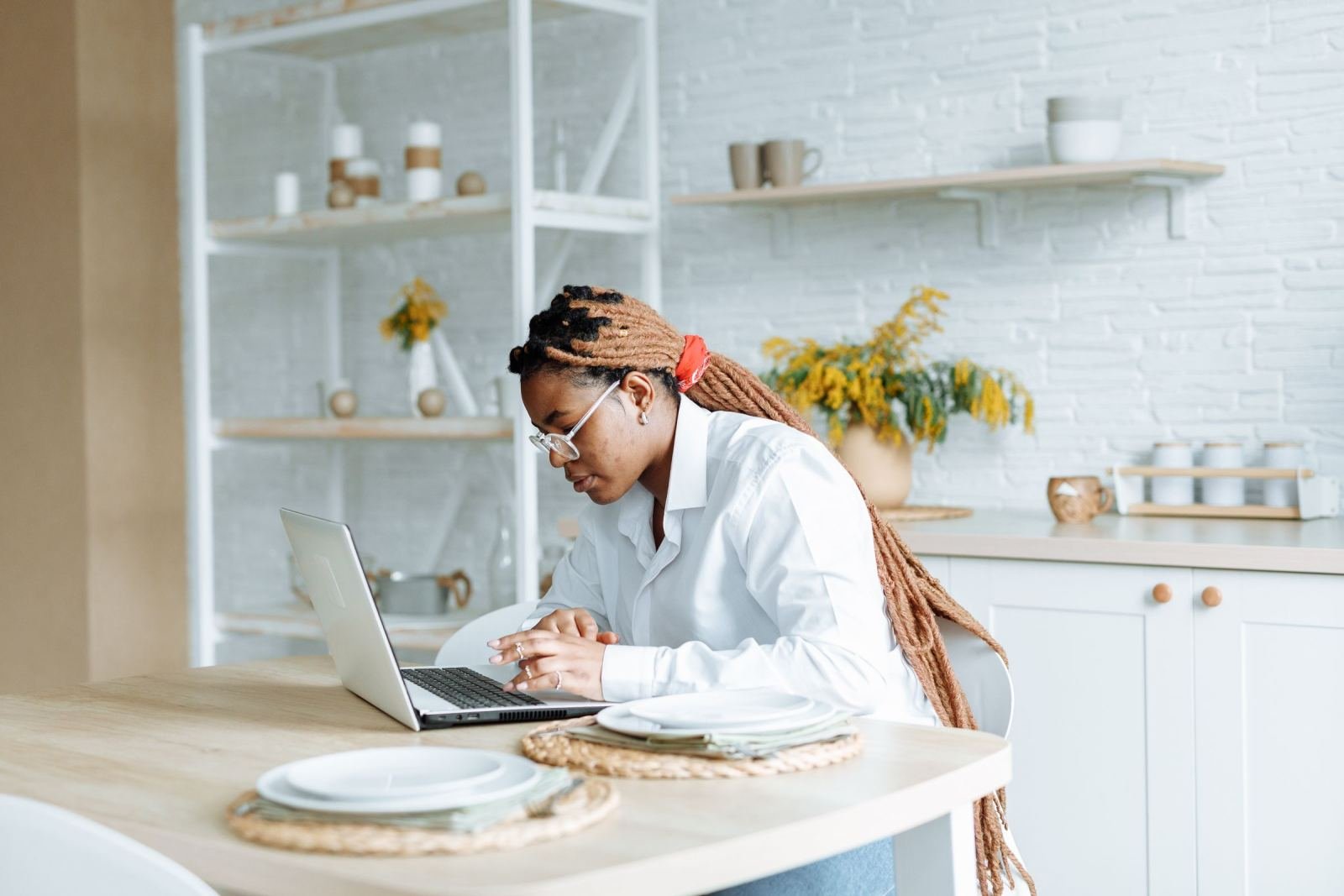 A woman with long braids and glasses uses a laptop at a bright kitchen table