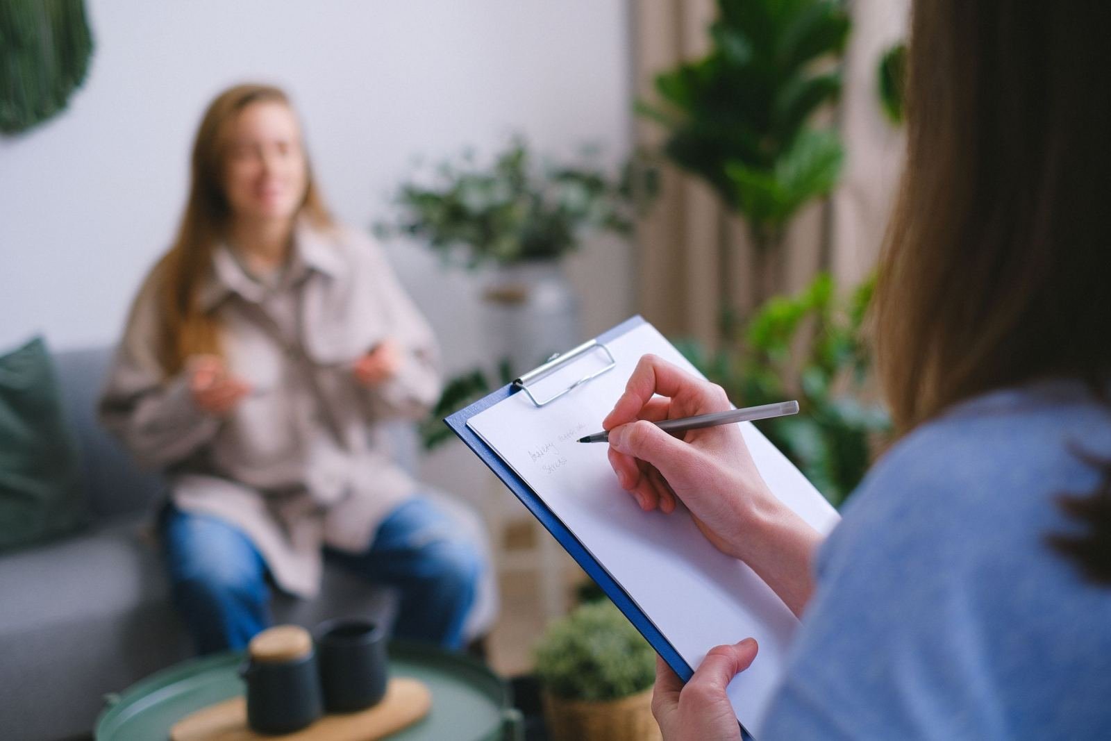 A clinician takes notes on a clipboard while a client speaks in the background
