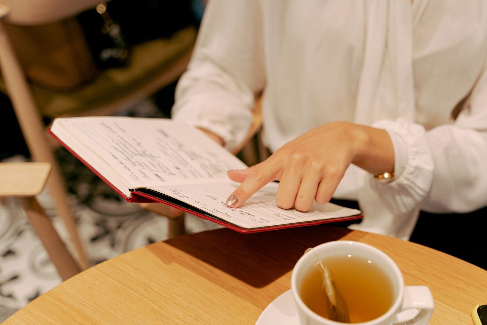 A woman in a white top shows notes in a red book beside a cup of tea.