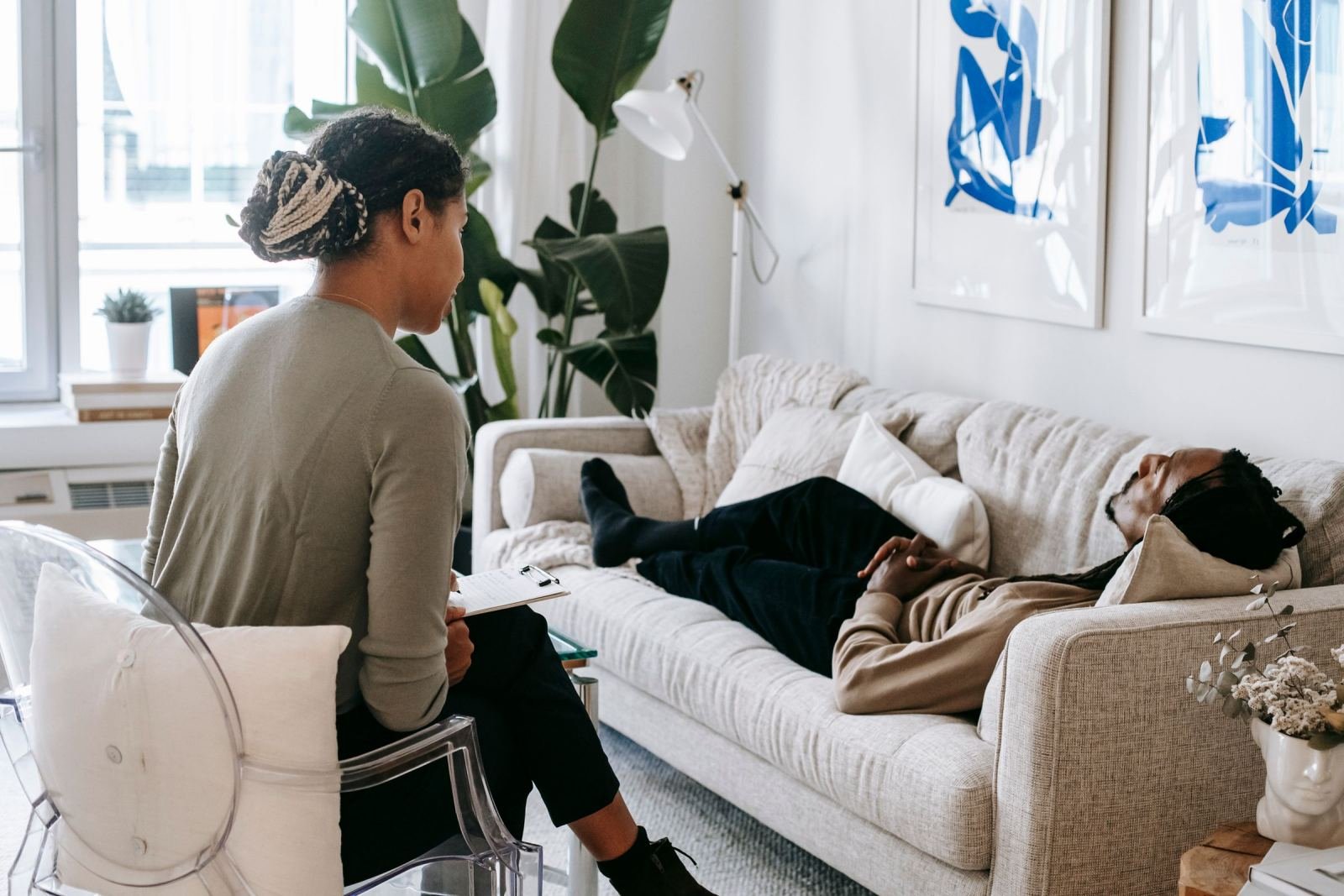 A counselor with a clipboard speaks to a person lying on a sofa in a bright office
