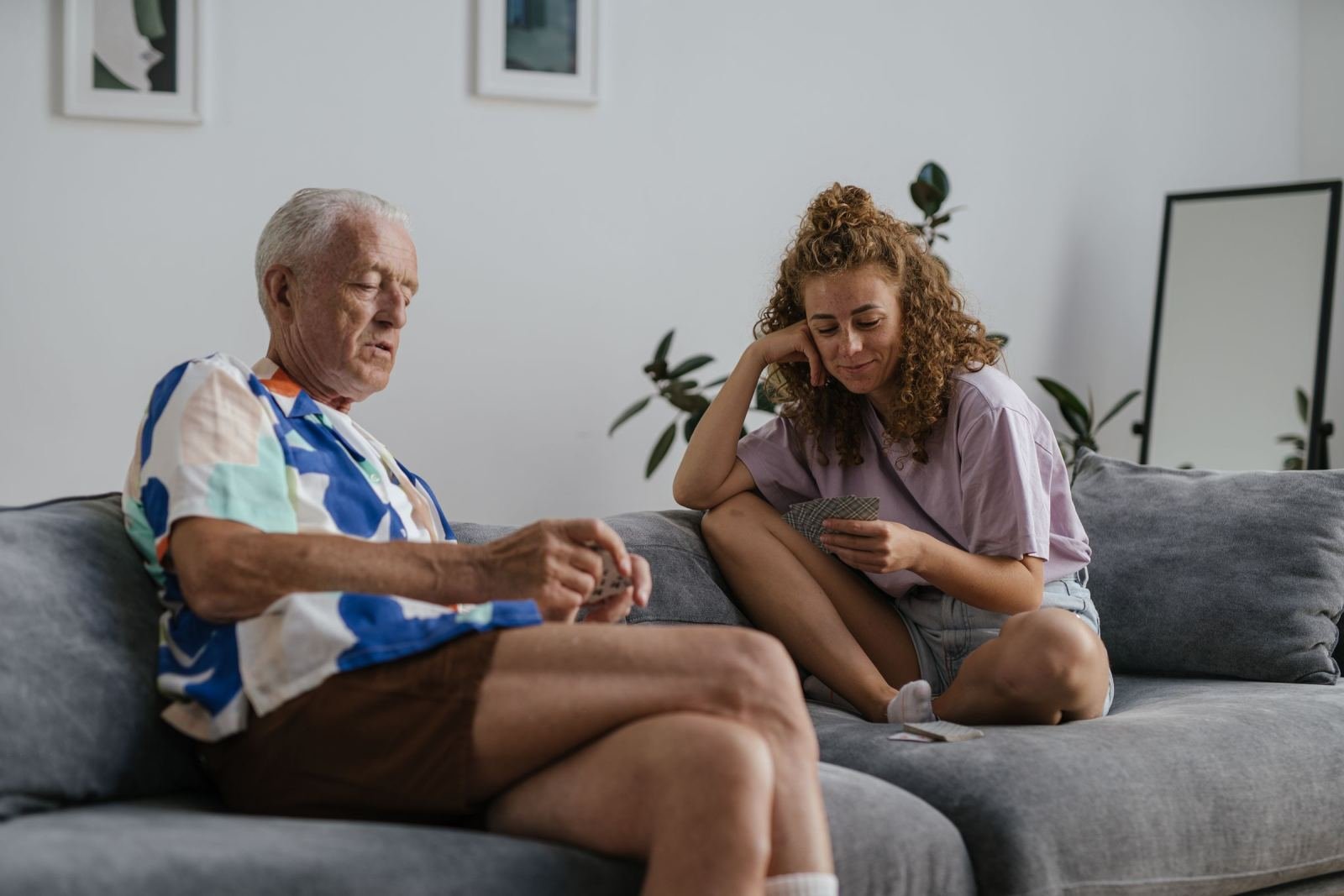 An older man and a younger woman play cards together on a gray sofa