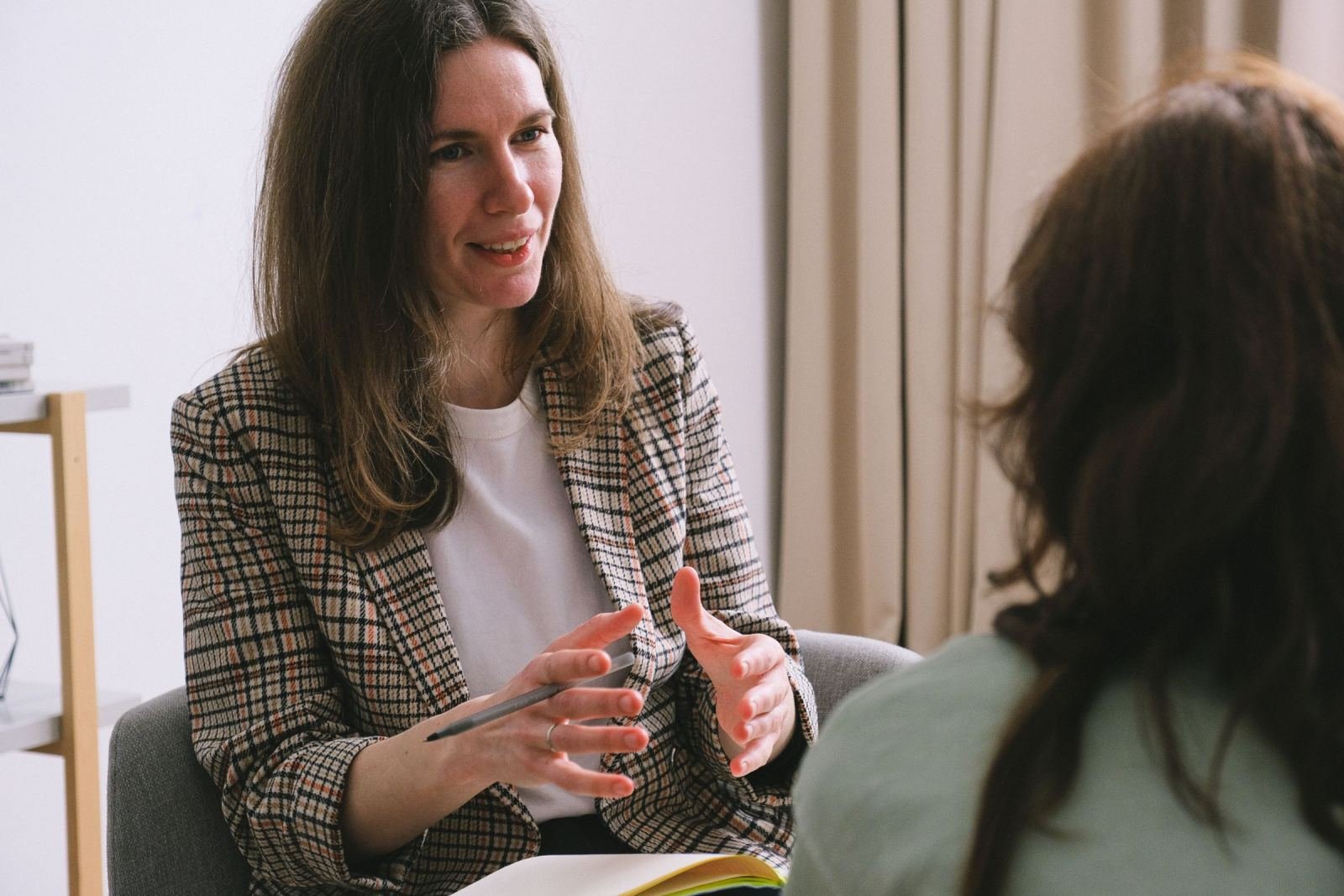 A woman in a plaid blazer gestures while speaking to another person in an office