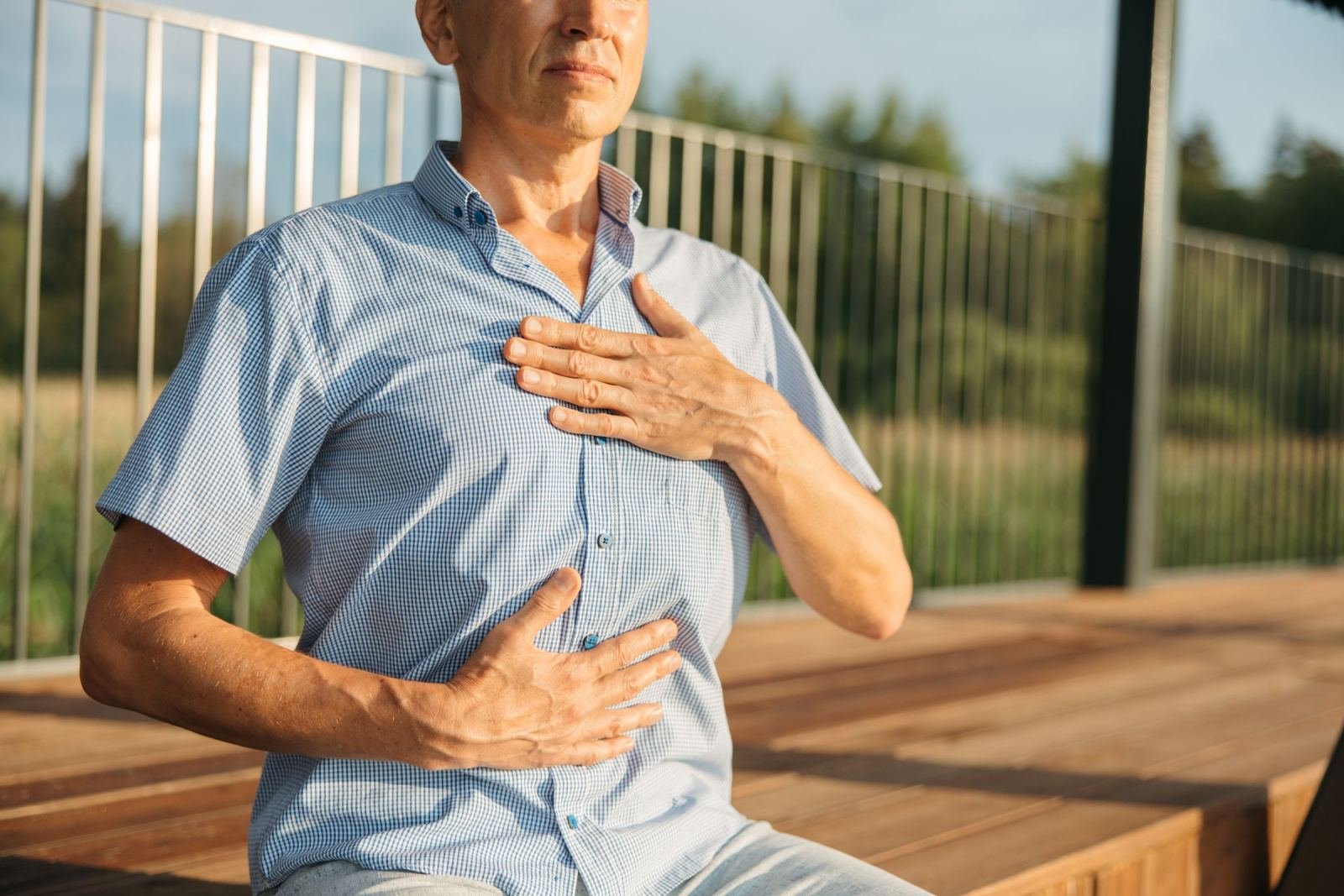 A man in a blue checkered shirt sits on a deck with his hands on his chest