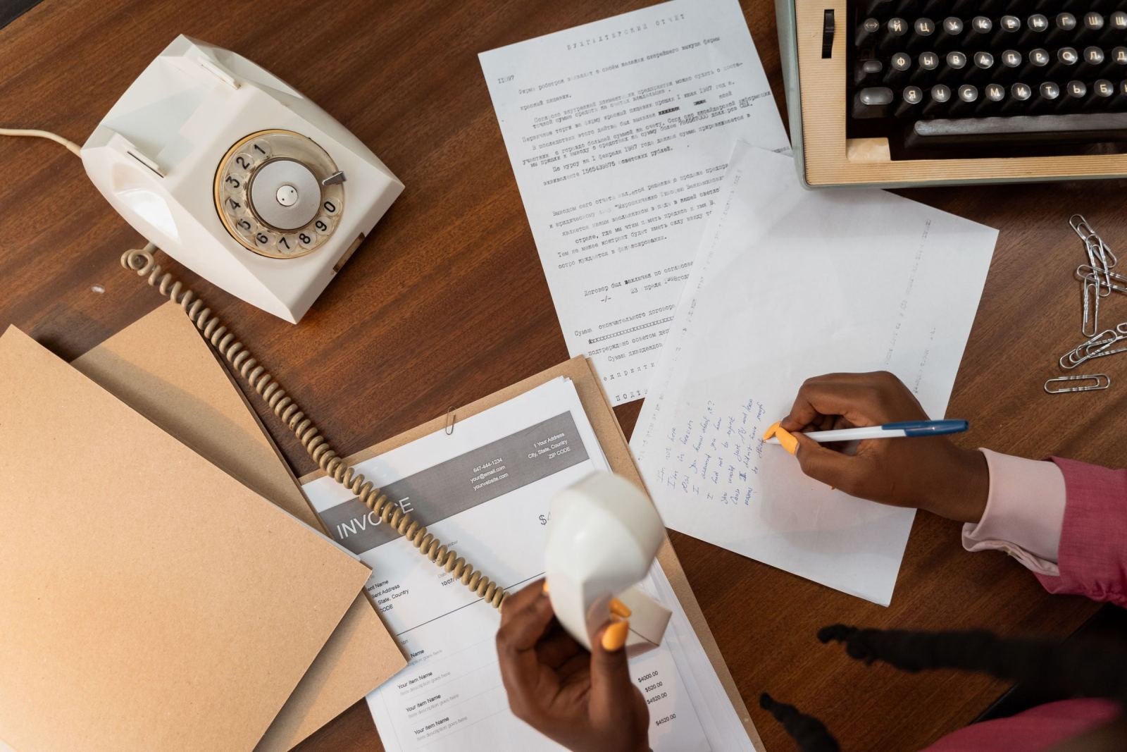 A person writes on a piece of paper at a desk with a rotary phone and typewriter