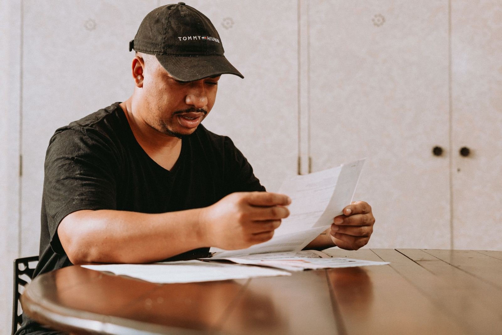 A man in a black cap reads a document at a wooden table
