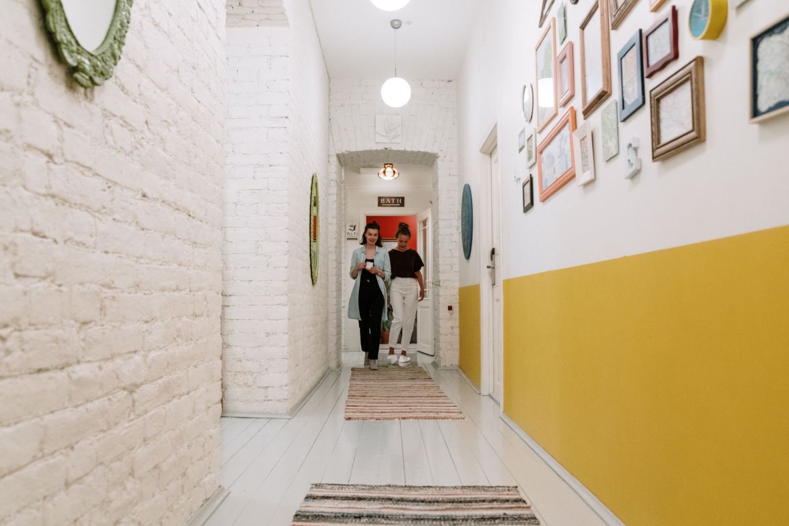Two women walk through a bright hallway with white brick walls and yellow paint