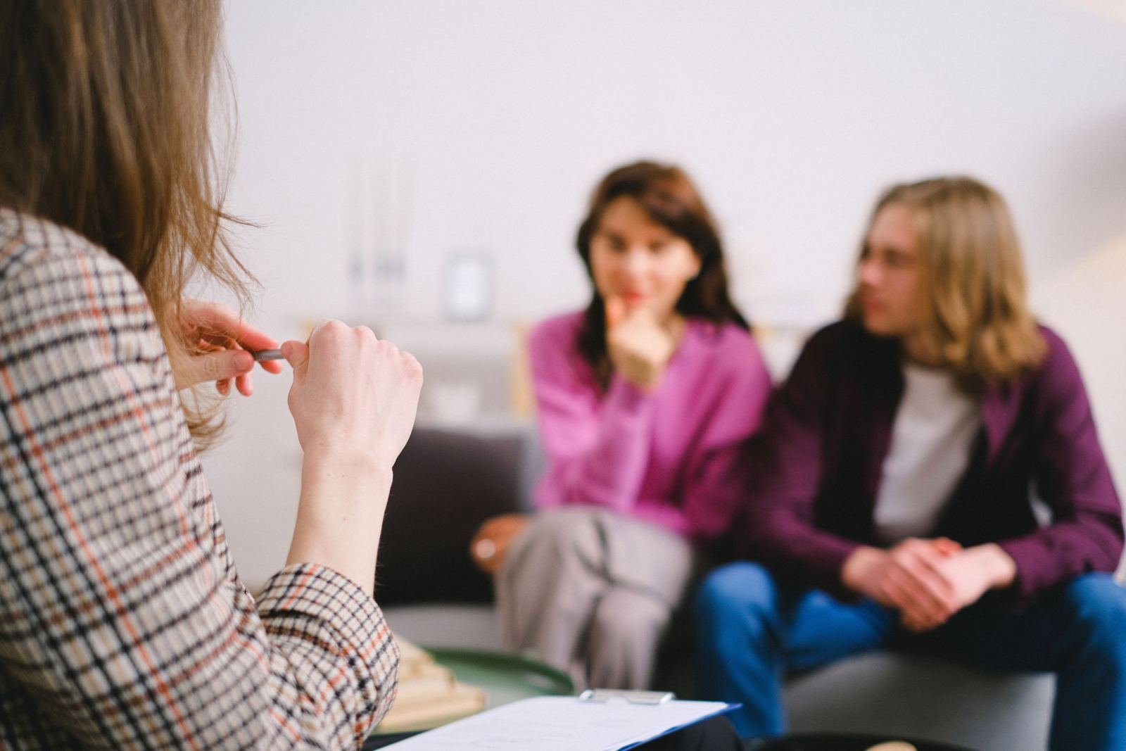 A specialist wearing a plaid jacket talks to a pair of people on a couch.