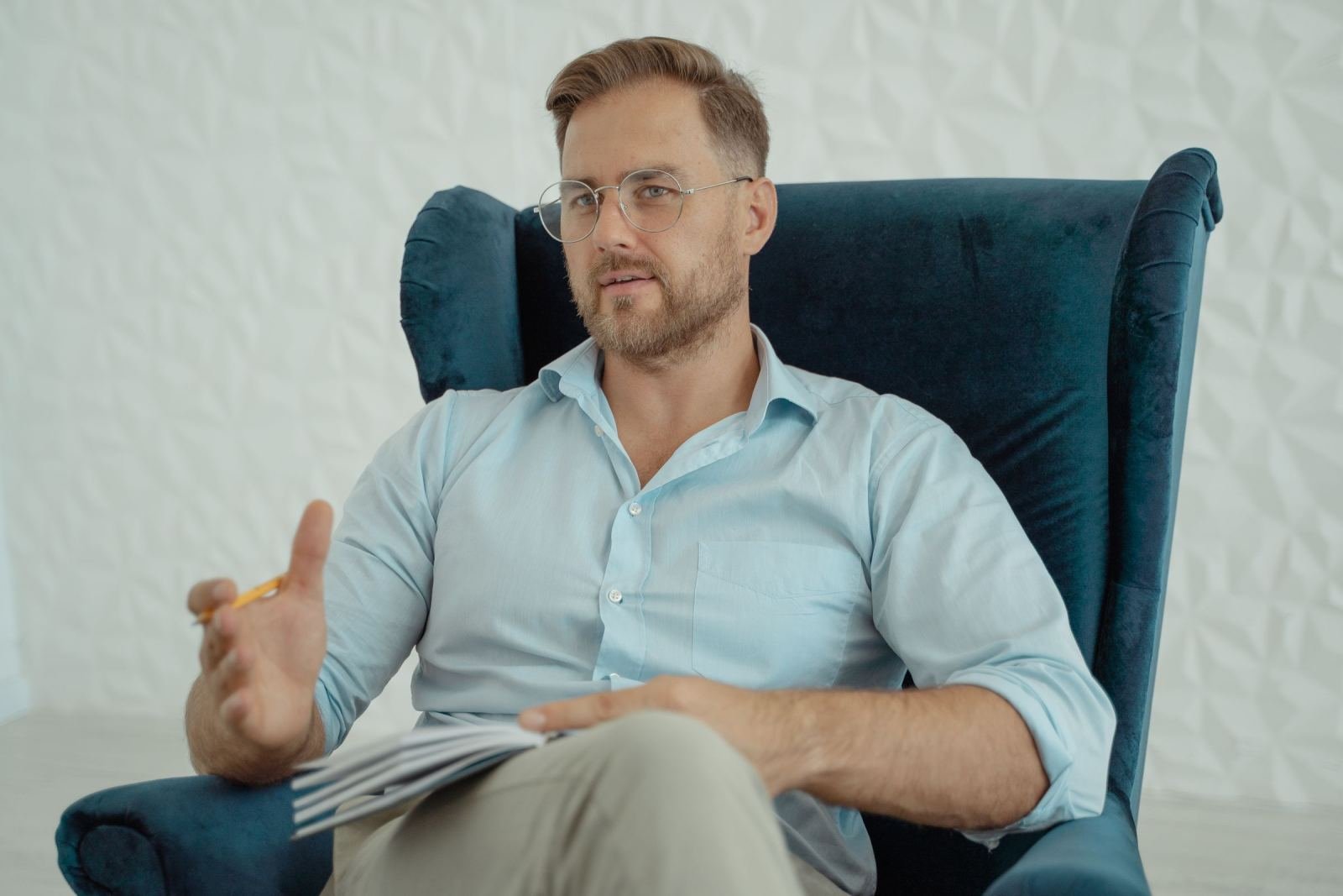 A man in a blue shirt and glasses sits in a blue armchair holding a notebook