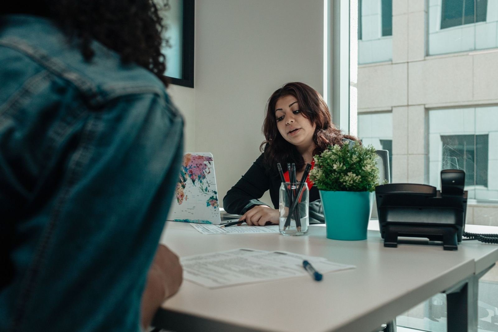 A woman sits at a desk and speaks with a person seen from behind