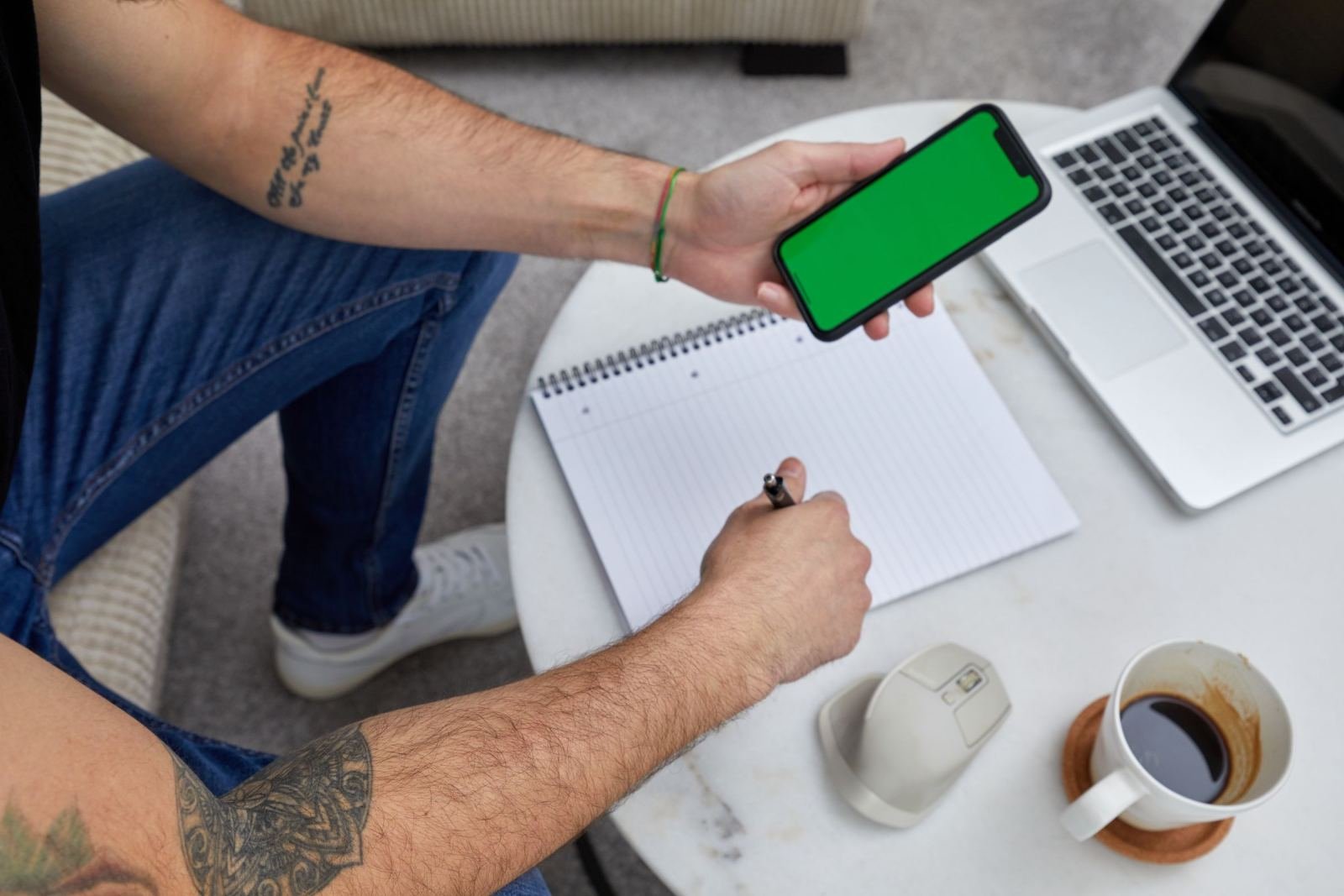 A person holding a smartphone and pen over a notebook at a table with a laptop and coffee