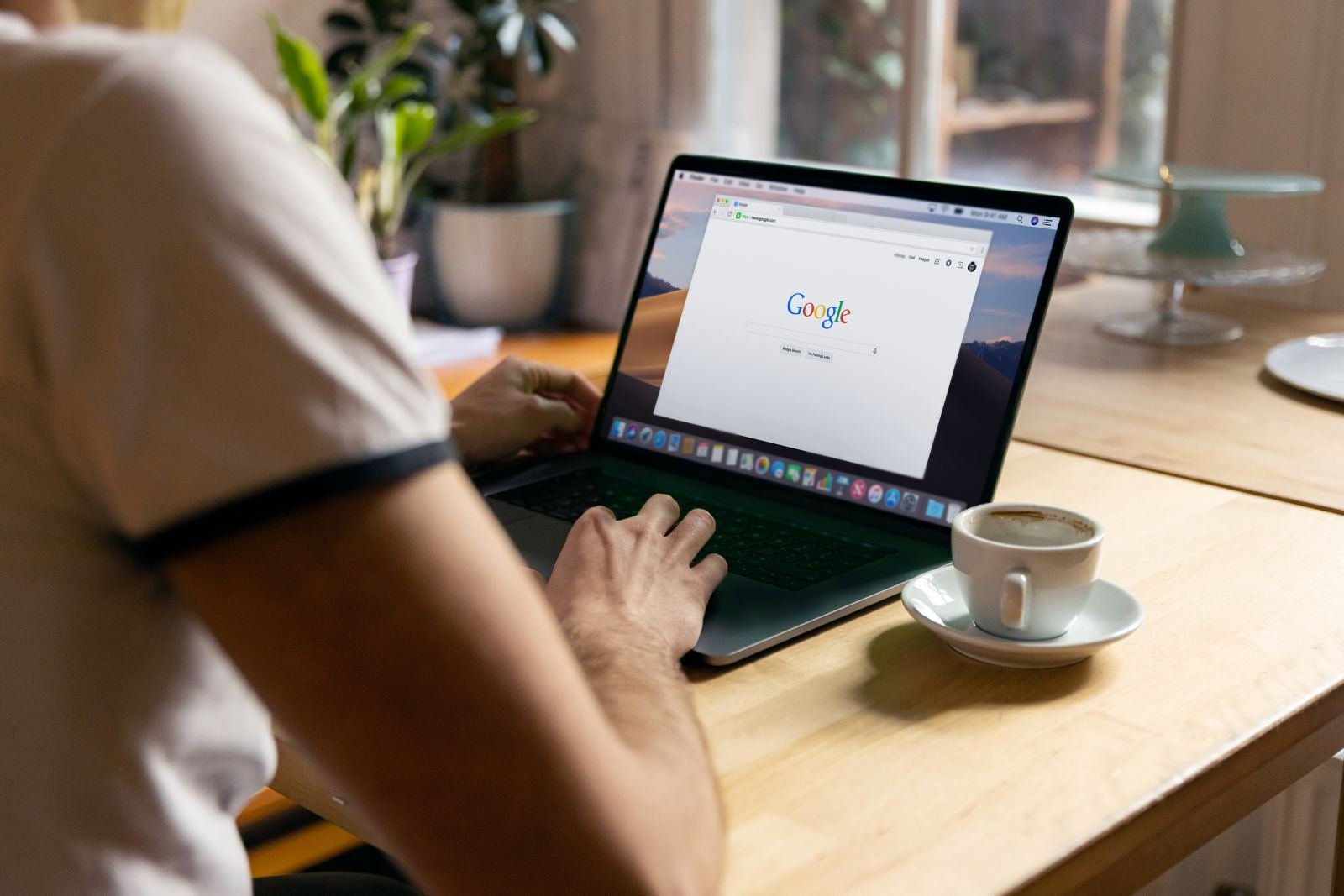 A person sits at a wooden table with coffee, using a laptop to browse the internet.