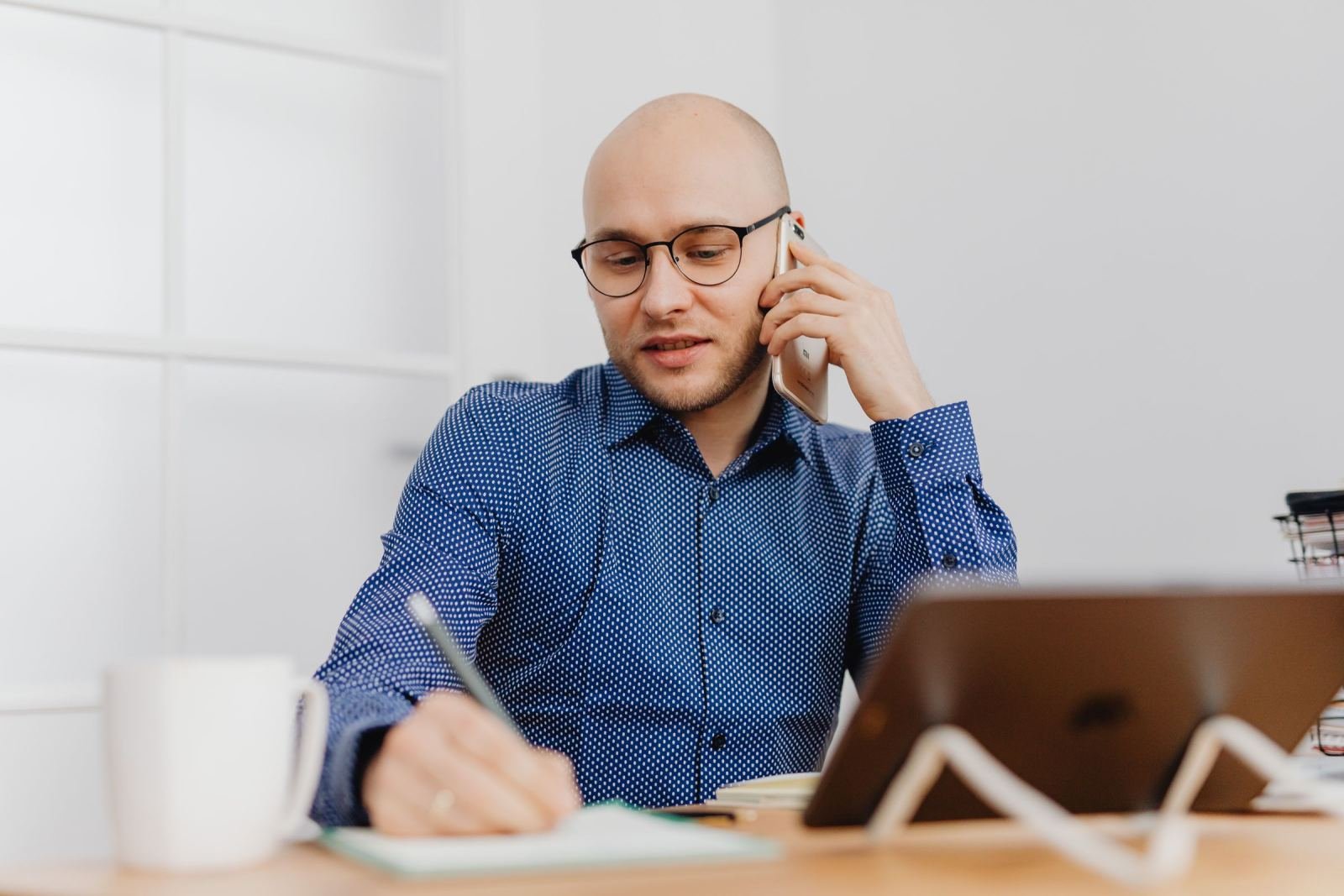 A man with glasses talks on a phone while taking notes at a desk
