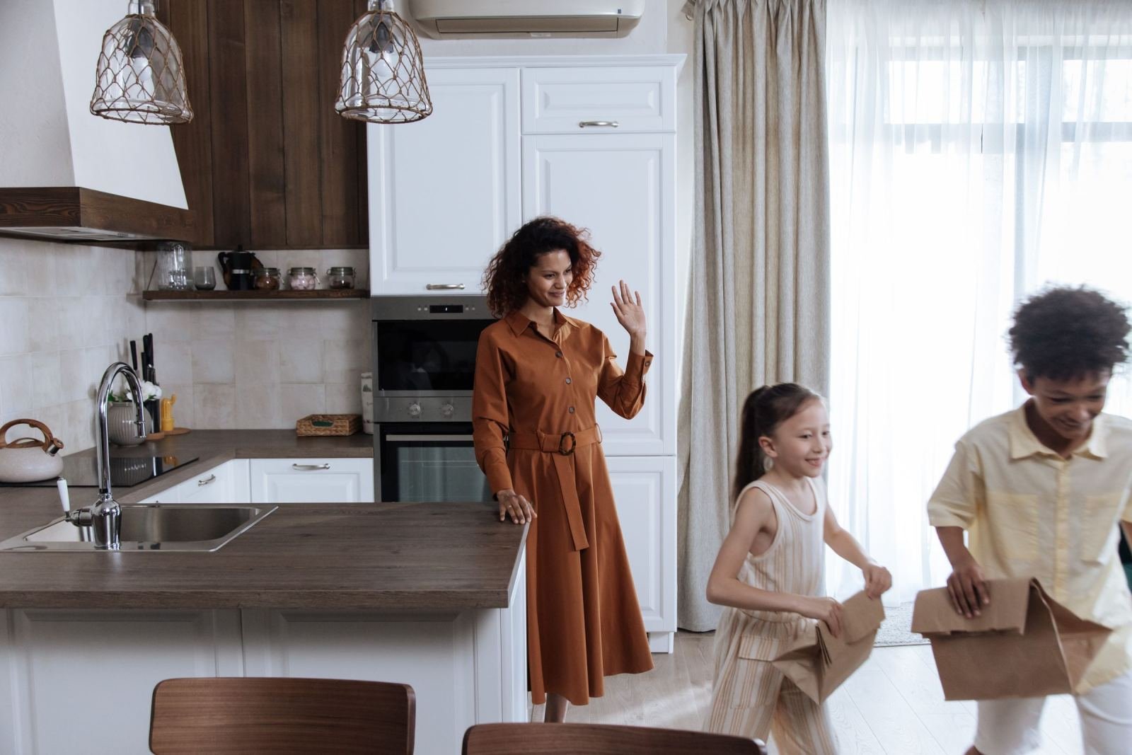 A woman waves to two children in a bright, modern kitchen