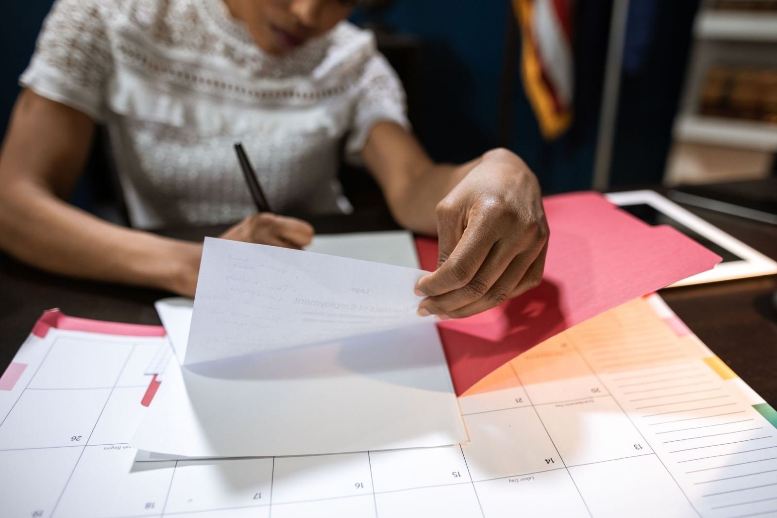 A person in a white lace top reviews a paper over a calendar at a desk